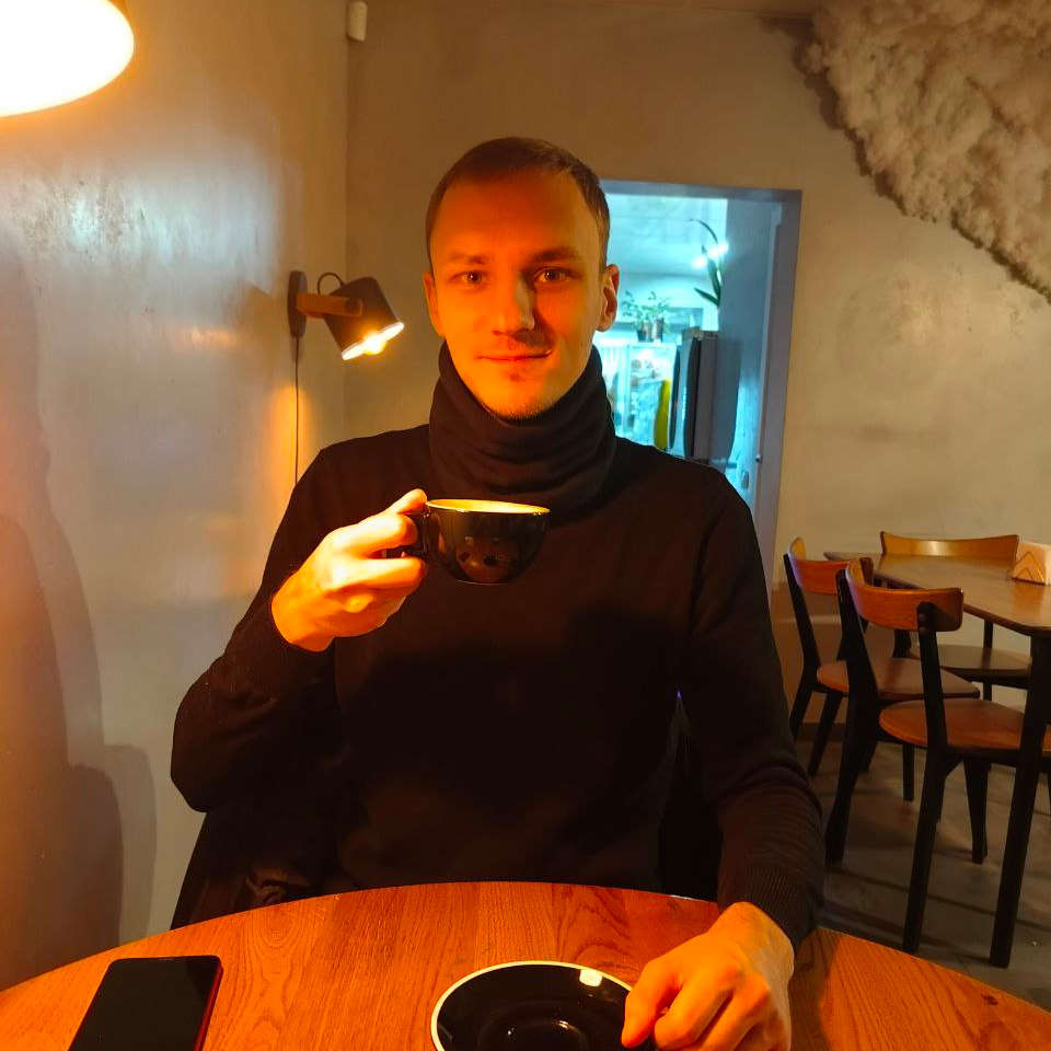 Хлопець 25 років із русявим волоссям і оливковими очима, в темному светрі за столиком тримає чашку кави.